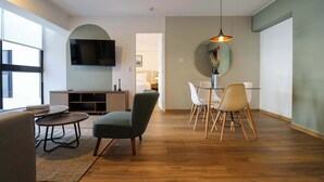 A modern living room with a flat-screen TV, a dining area with a glass table and chairs, and a large window providing natural light.