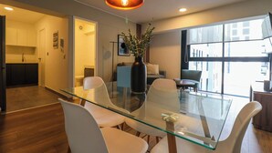 A modern dining area with a glass table and white chairs, a vase with greenery, and a large window with a city view.