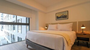 A modern hotel room with a large bed, a nightstand, a lamp, and a view of a building through a glass balcony.