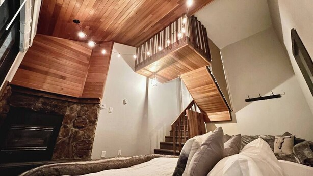 A bedroom with a stone fireplace, wooden ceiling, and a staircase leading to an upper level. - Small Cabin, Big Comfort. Ski-in/Ski-Out, Hike, Bike & Chill. Hot Tub & Sauna. (Kimberley)