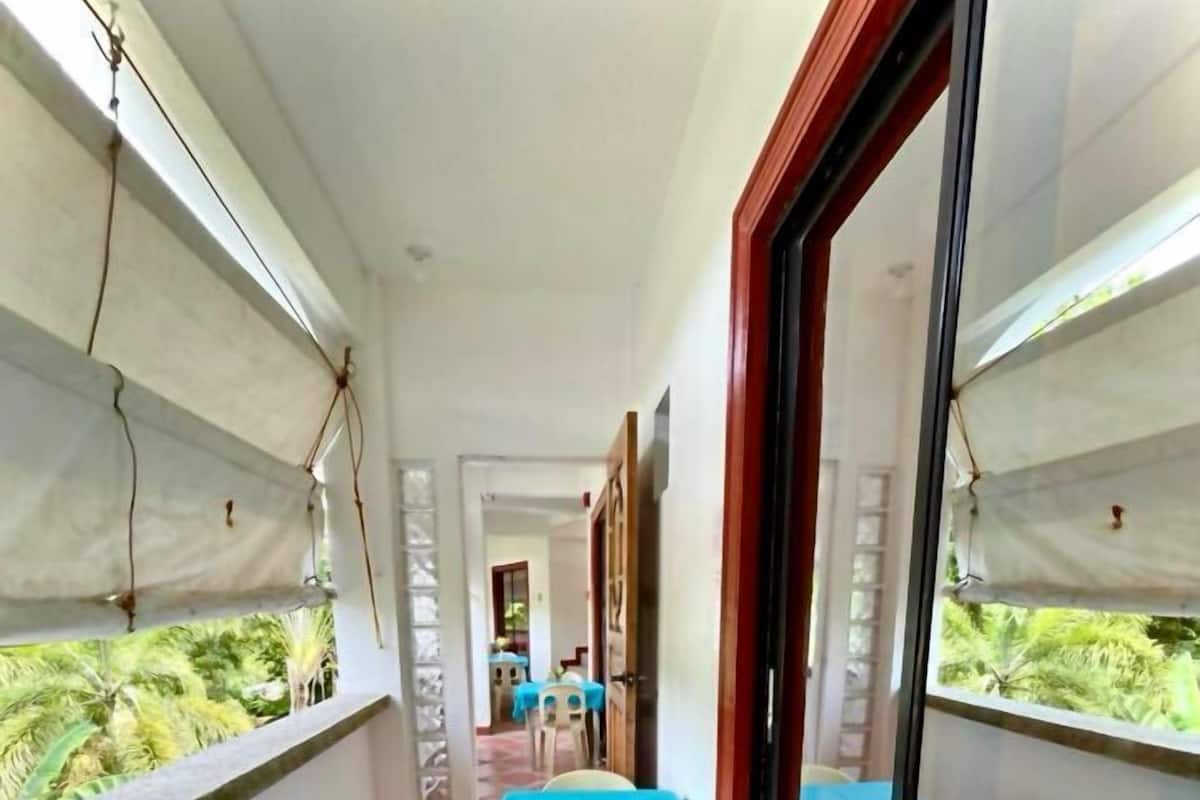 Varanda com mesa azul e duas cadeiras de plástico, porta de vidro e vista para vegetação.