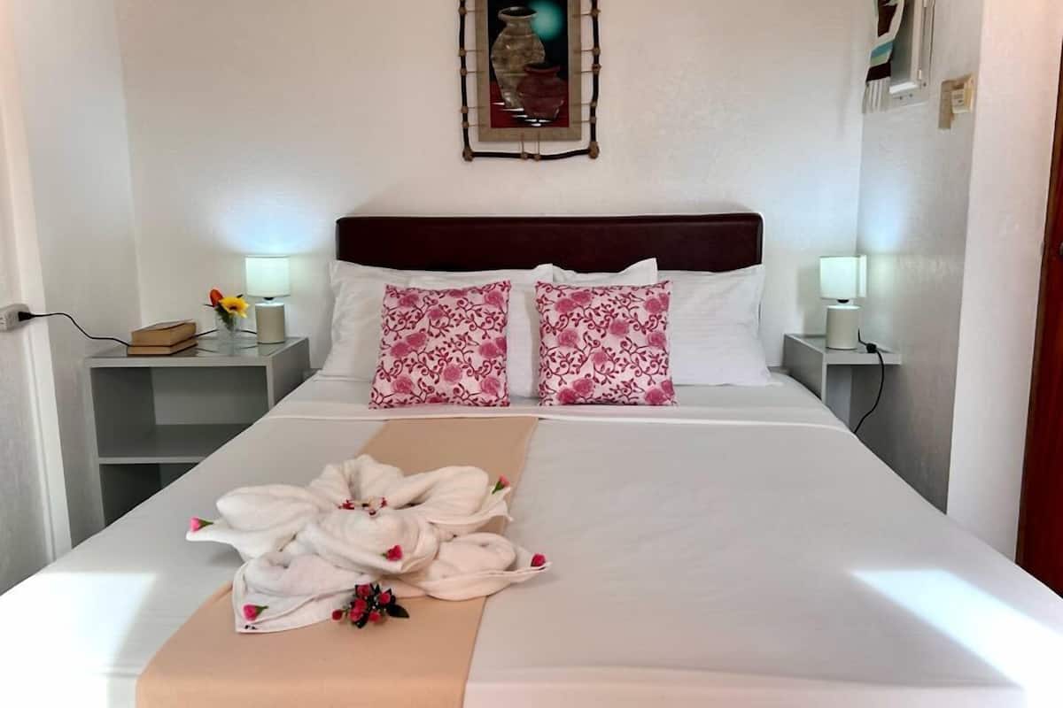 Cama bem arrumada com almofadas florais rosa, mesa de cabeceira com abajur e moldura de quadro na parede.