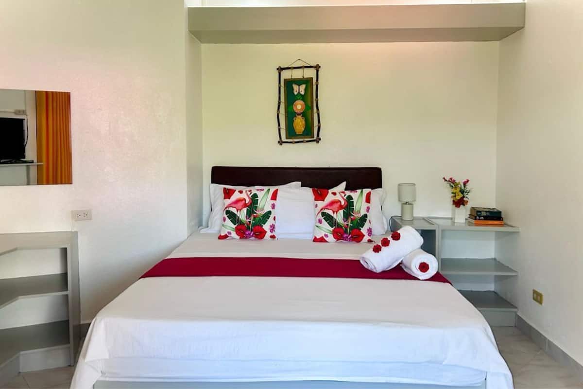 Uma cama bem arrumada com almofadas florais, um cobre-leito vermelho e uma toalha enrolada ao lado.