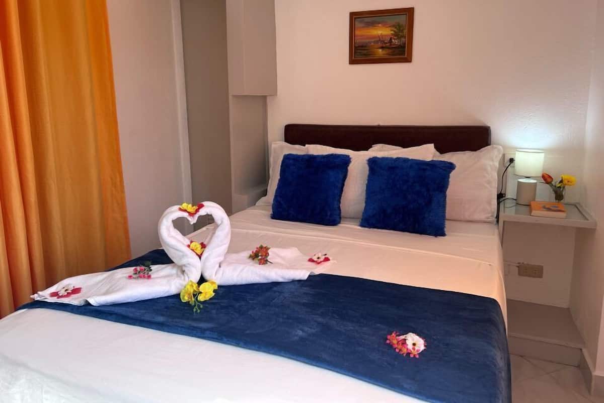 Quarto de hotel com cama bem arrumada, um quadro na parede e uma mesa de cabeceira com luminária e flores.
