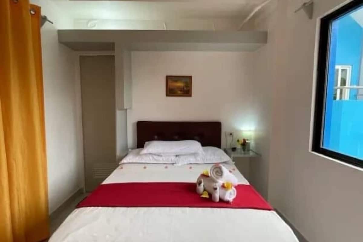Quarto de hotel com uma cama, um cobertor vermelho, uma mesa pequena com luminária e uma janela com vista para edifícios.