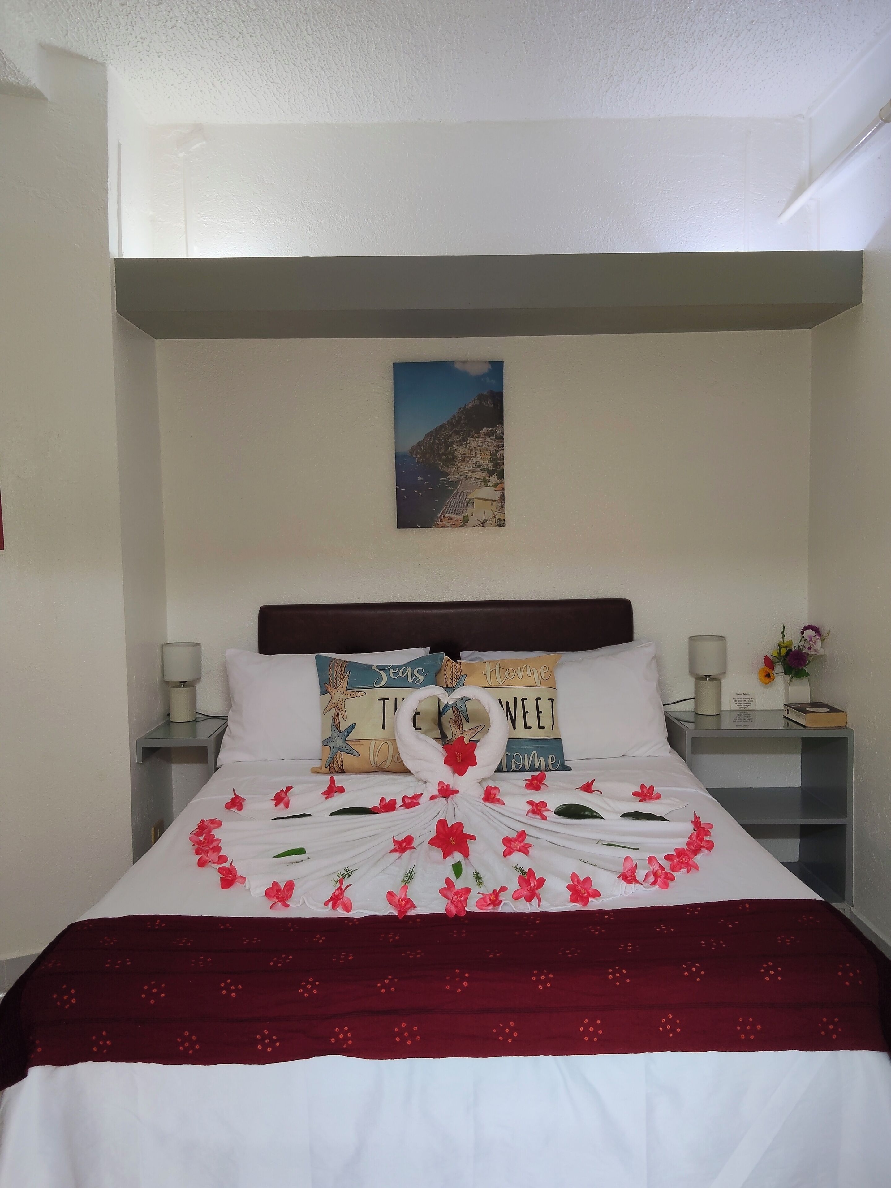 Una cama bien hecha con una toalla dispuesta en forma de corazón, un cuadro enmarcado en la pared y dos mesitas de noche con lámparas y un pequeño jarrón de flores.