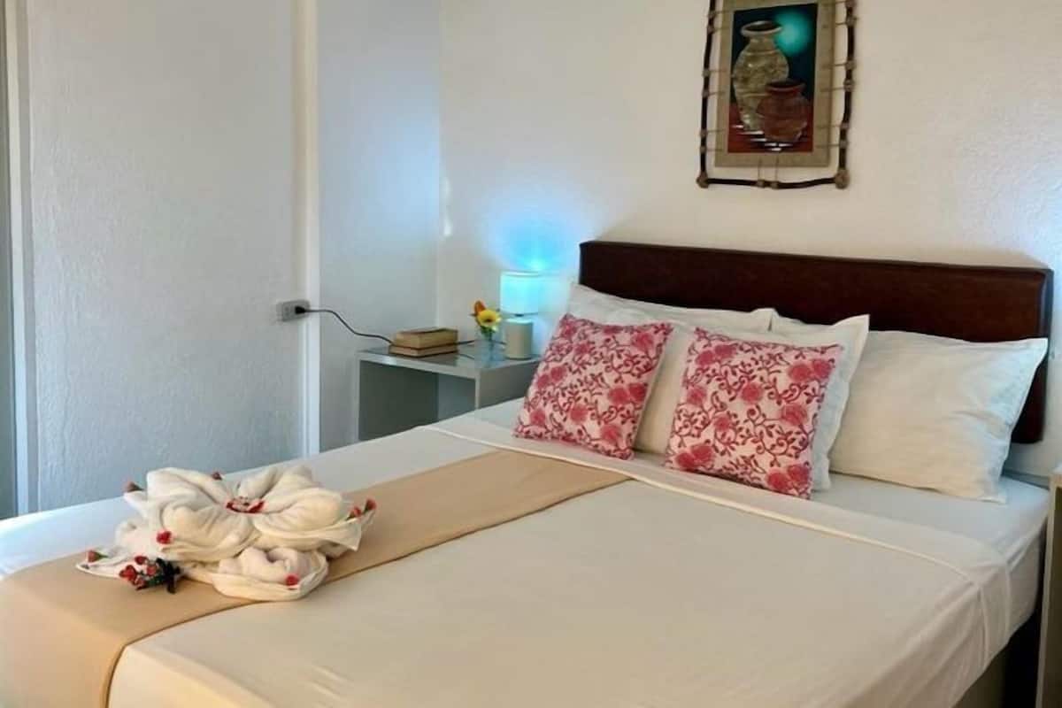 Uma cama bem arrumada com almofadas florais, uma luminária de cabeceira e um quadro na parede.