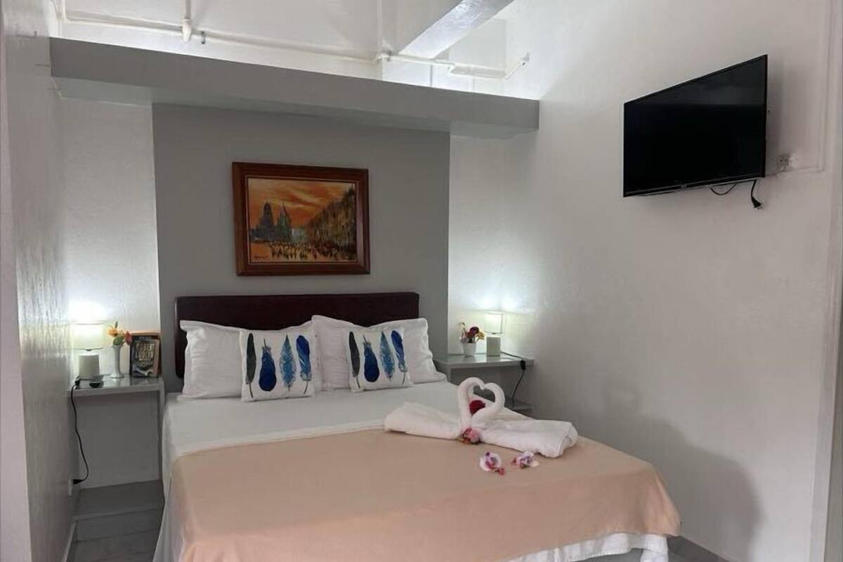 Quarto de hotel com uma cama, um quadro na parede, uma TV fixada na parede e uma mesa de cabeceira com abajur e um livro.