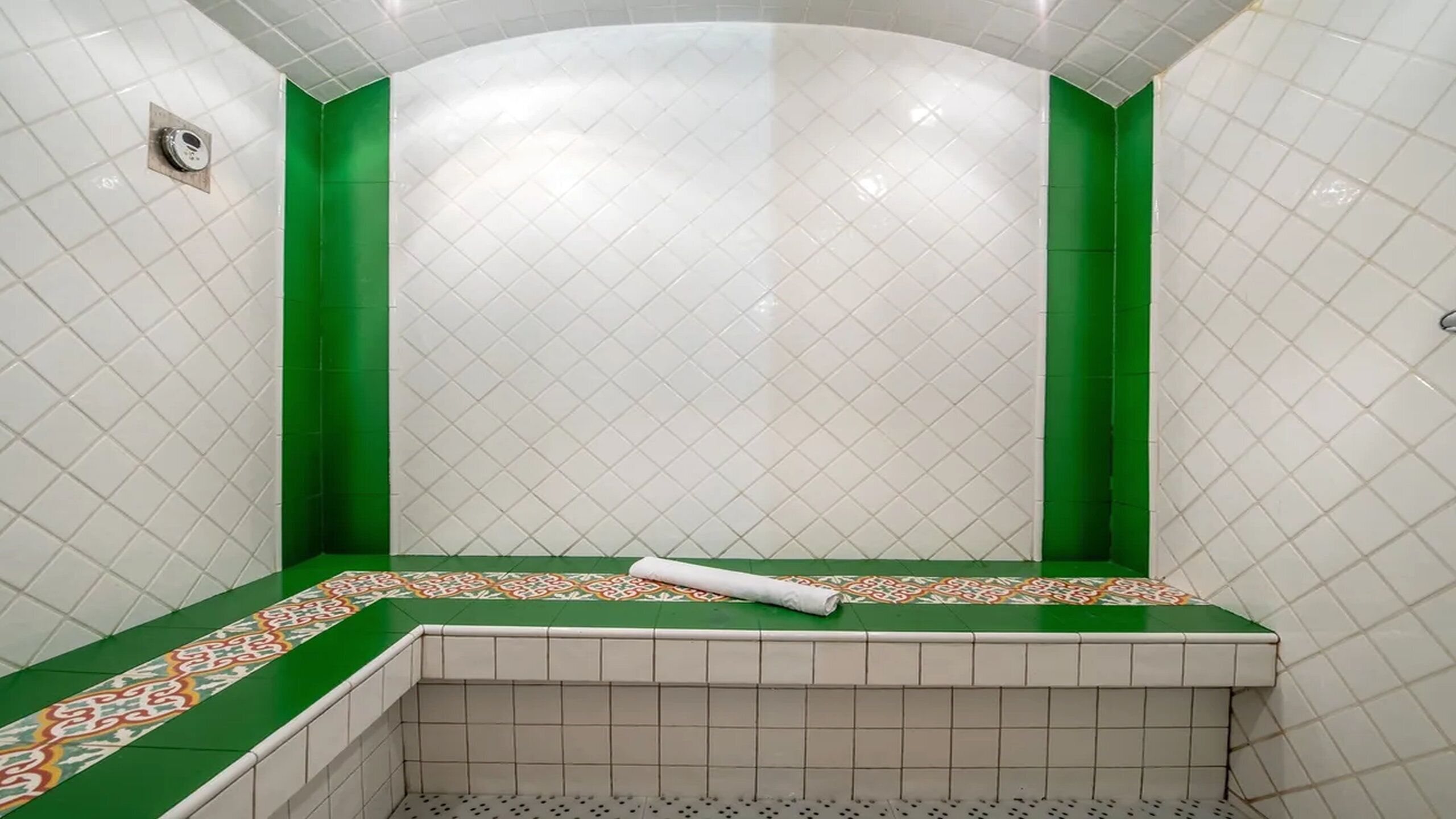 A bathroom with white and green tiles, a shower area, and a towel on the floor.