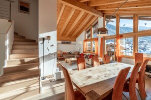 A spacious living area with a wooden staircase, a dining table, and large windows offering a view of the mountains. - Fantastique mansarde appartement (Zermatt)
