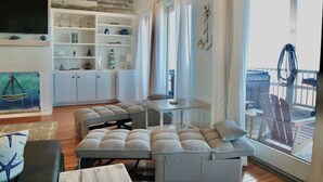 A modern living room with a large TV, a built-in shelf unit, and a balcony with outdoor seating. - Family Beachfront 5BR Home On Dauphin Island, Hot Tub! (Dauphin Island)