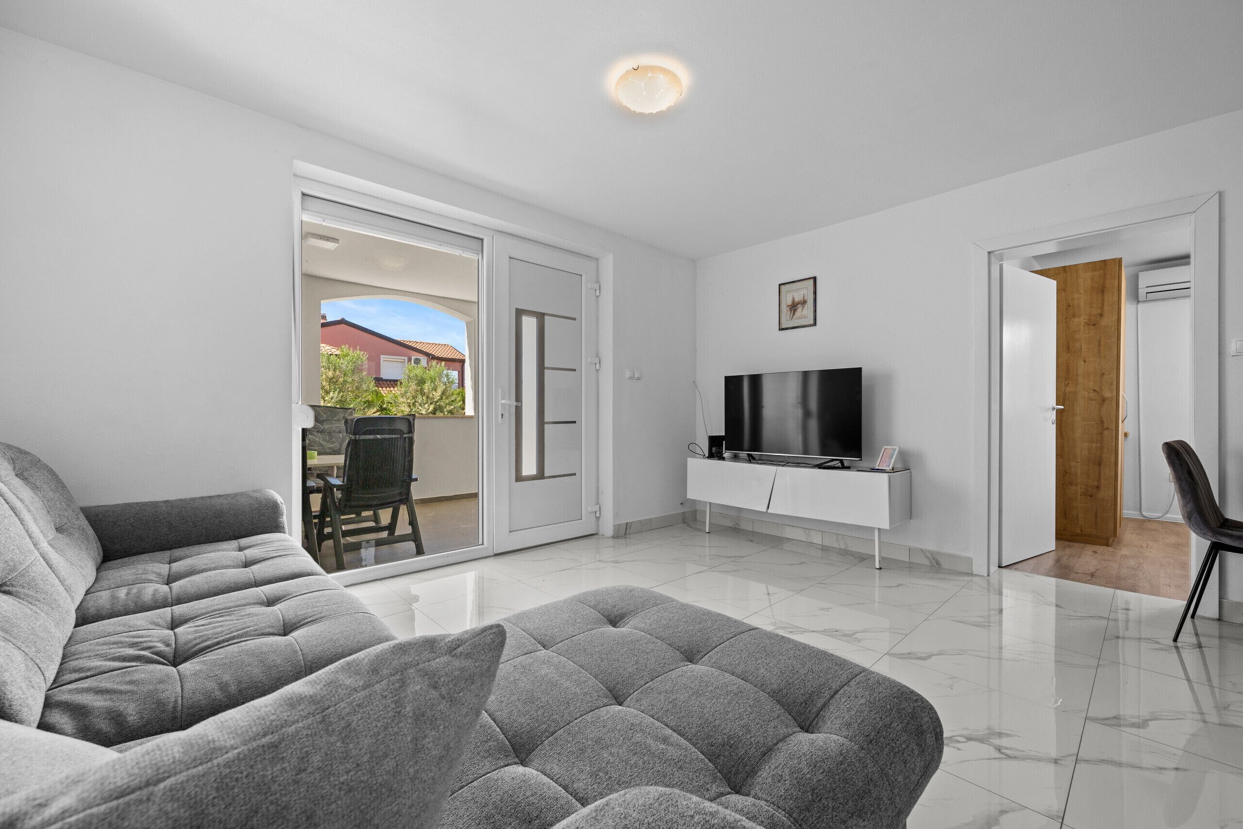 Un salon moderne avec un canapé gris, un téléviseur à écran plat posé sur un meuble blanc et une porte-fenêtre coulissante donnant sur un espace extérieur.
