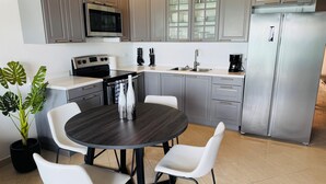 A modern kitchen with grey cabinets, a stainless steel refrigerator, a microwave, and a coffee maker. There is a dining table with four white chairs and a potted plant. - Beachfront Condo on Boardwalk – 2BR/2BA with Views (Philipsburg)