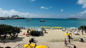 A beach with yellow umbrellas, lounge chairs, and a view of boats in the water. - Beachfront Condo on Boardwalk – 2BR/2BA with Views (Philipsburg)