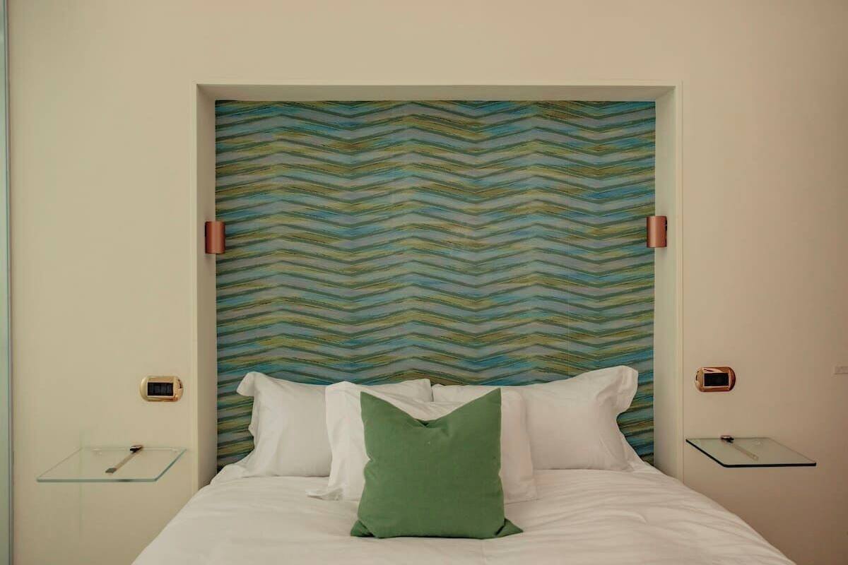 A hotel room with a bed, a green pillow, a striped wall, and a small bedside table with a lamp.