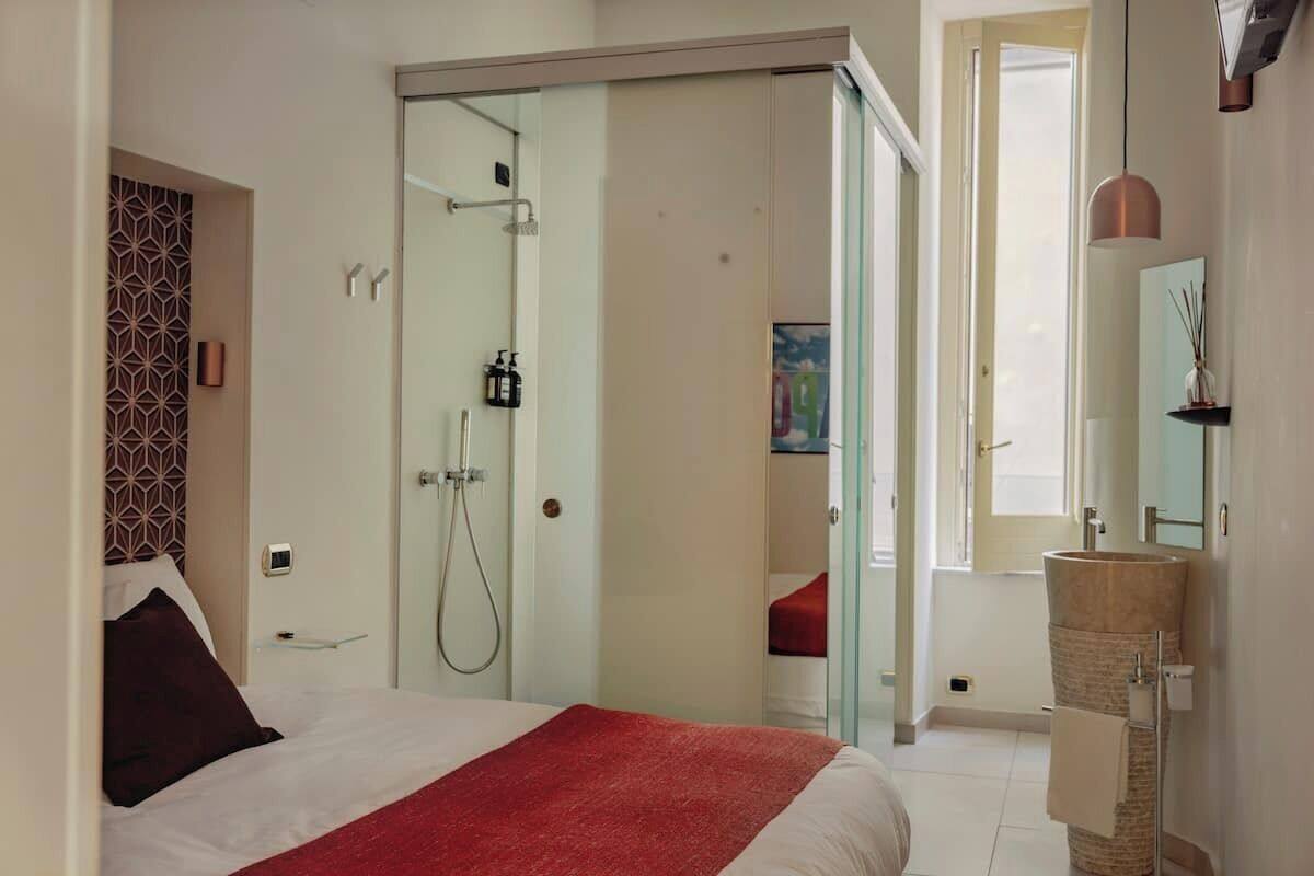A modern hotel room with a bed, a glass shower enclosure, a sink, and a large window.
