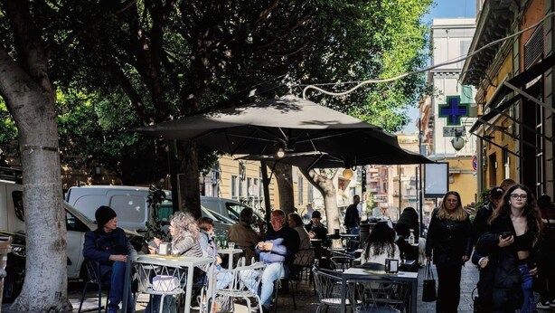 An outdoor café with tables and chairs under a large umbrella, surrounded by trees and buildings. - Da Celeste (Naples)