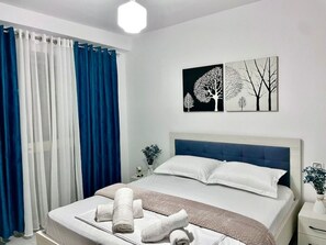 A neatly made bed with white linens and a blue headboard, a bedside table with a vase of flowers, and two wall art pieces depicting trees.