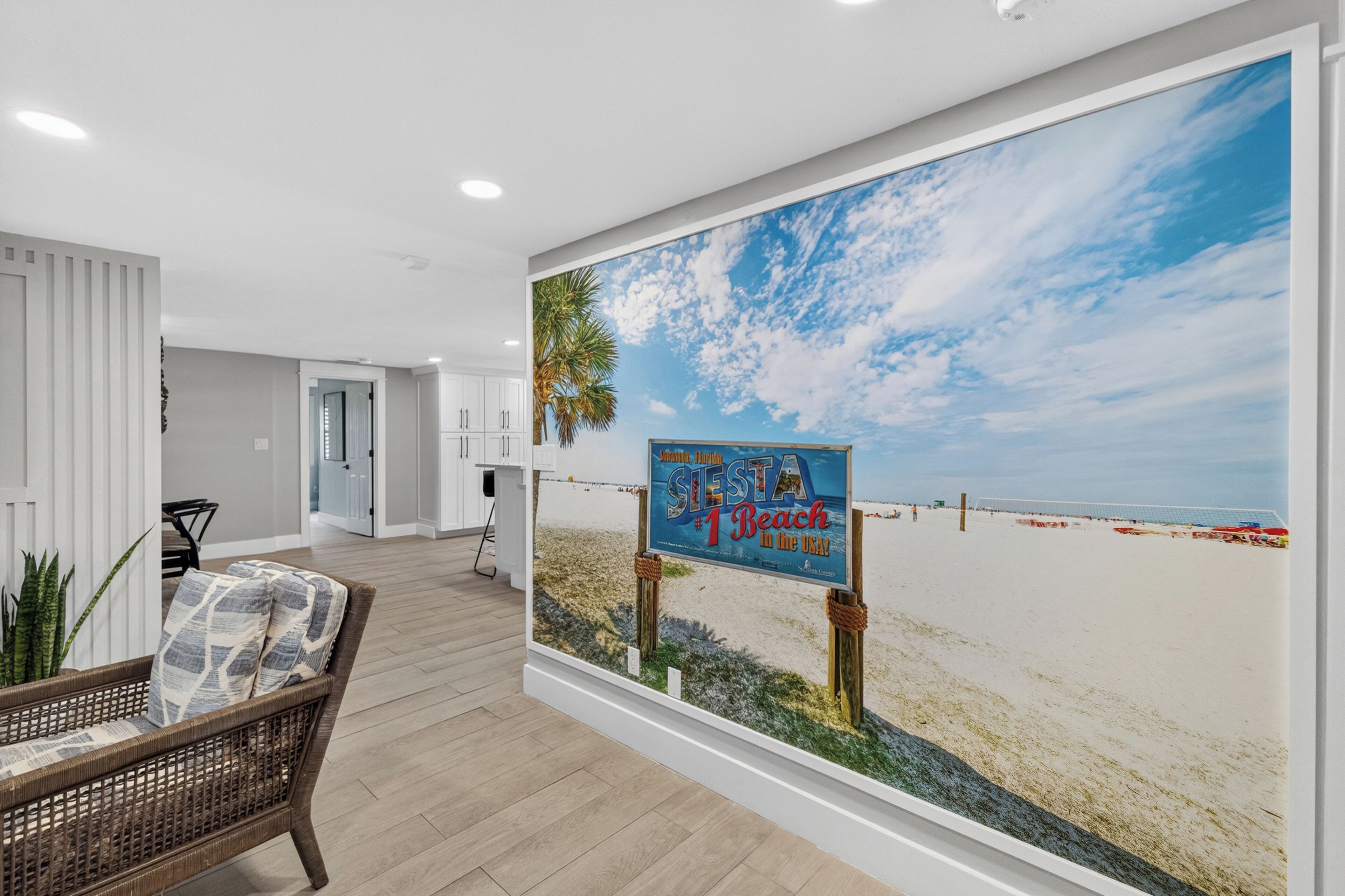 Een muurschildering met een strandthema, met een bord waarop staat 'Siesta #1 Beach in the USA', in een kamer met een rieten stoel en uitzicht op een deur die leidt naar een andere kamer.