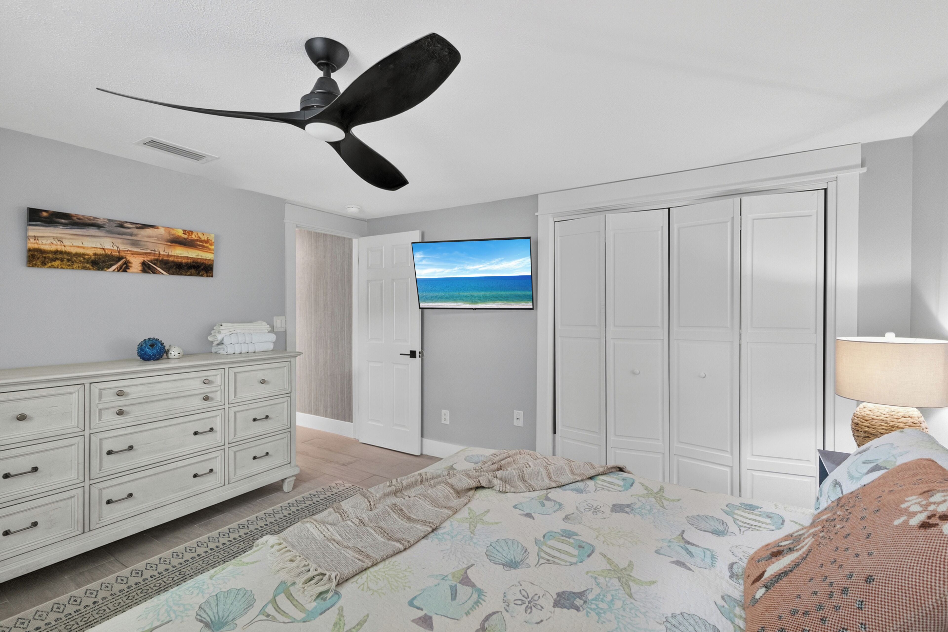 Ein Schlafzimmer mit Bett, Kommode, Deckenventilator, einer Tür zu einem anderen Raum und einem an der Wand befestigten Fernseher.