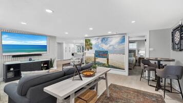 A modern living room with a large flat-screen TV, a grey sofa, a coffee table, and a painting of a beach scene.