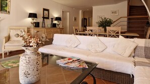 A modern living room with a white sofa, a glass coffee table, and a wicker chair. There is a large vase with flowers on the floor, and a staircase is visible in the background. - Beautiful private villa for 6 people with pool, WIFI, TV and patio (Praia Da Falesia)