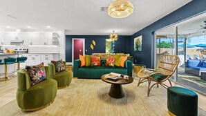 Ein modernes Wohnzimmer mit einem grünen Sofa, zwei Sesseln, einem Couchtisch und einem Essbereich, im Hintergrund eine weiße Küche.