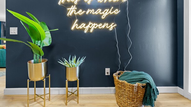 Ein Zimmer mit einer dunklen Wand, zwei goldfarbene Pflanzenständer mit Grünpflanzen und eine Neonreklame mit der Aufschrift 'This is where the magic happens'.