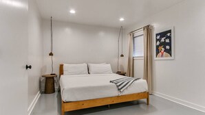 Une chambre à coucher au style minimaliste, avec un lit en bois, du linge de lit blanc, une petite table de chevet et un seul luminaire fixé au mur.