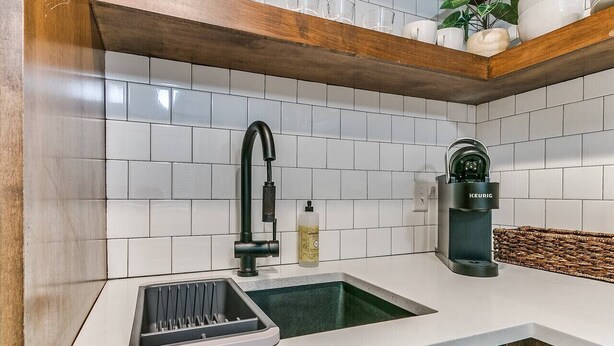 Une cuisine avec des carreaux blancs, un robinet noir, une machine à café et un évier équipé d’un égouttoir à vaisselle.