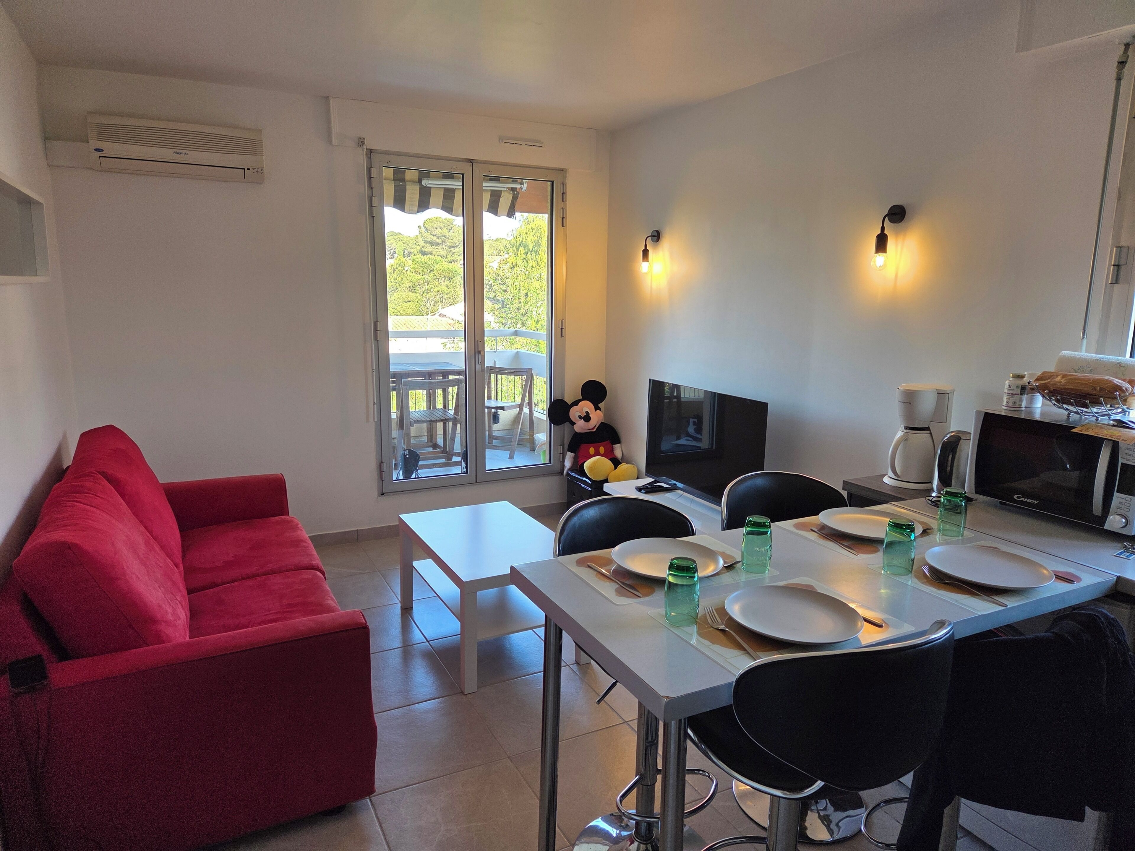 A compact hotel room with a dining area, a red sofa, a television, and a Mickey Mouse plush toy.