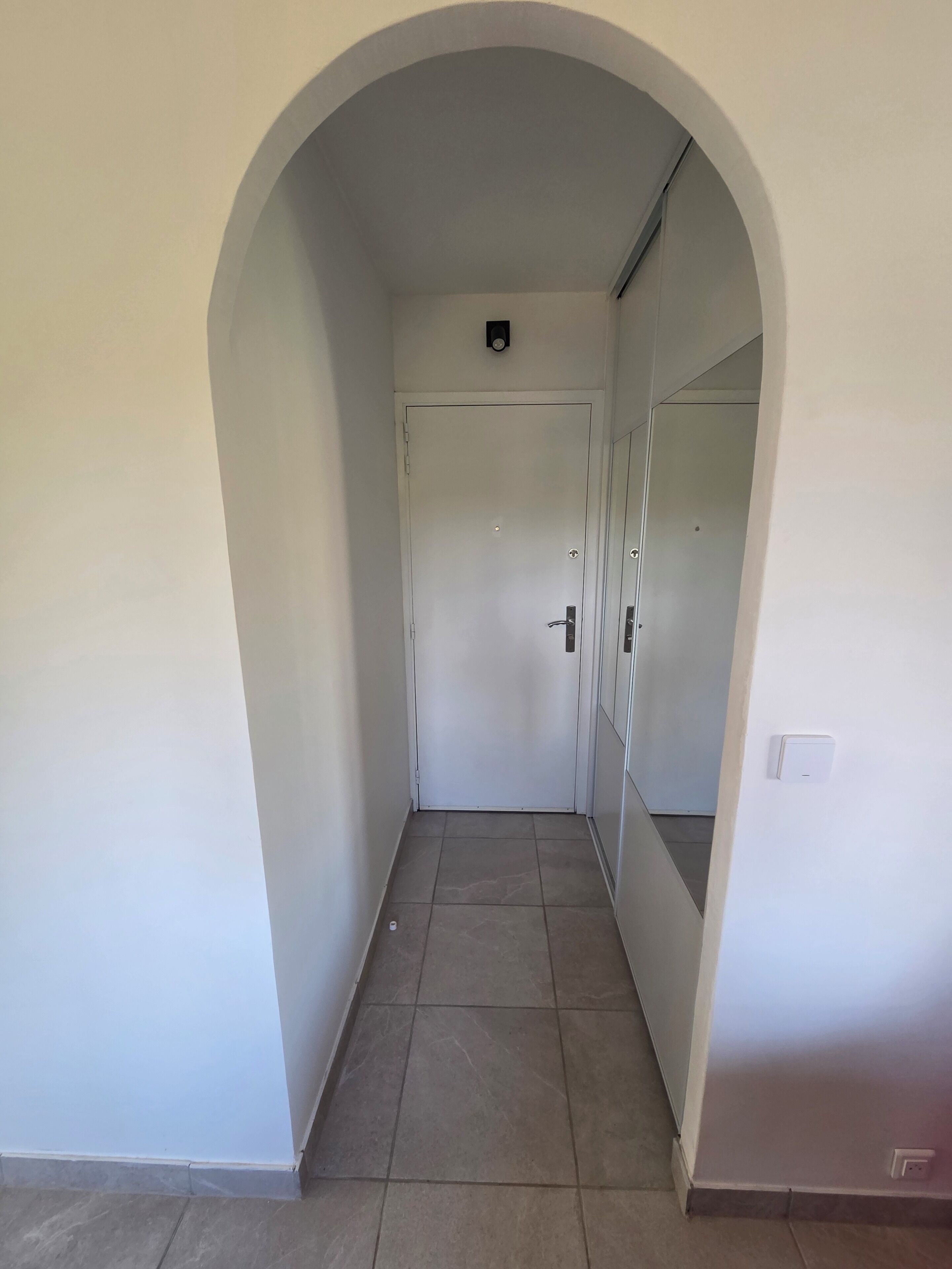 A hallway with a white door, a tiled floor, and a white wall with a light switch.