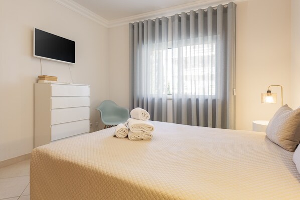 A neatly made bed with a quilted cover, a flat-screen TV mounted on the wall, a chair, and a window with curtains.