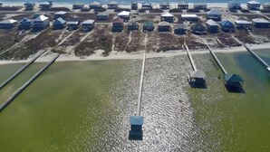 Vista aérea de una zona costera con varias construcciones pequeñas, probablemente cabañas o casas, dispuestas en filas a lo largo de la orilla. Se observan dos canales o muelles importantes que se adentran en el agua.