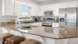 Una cocina moderna con armarios blancos, electrodomésticos de acero inoxidable, una encimera de granito y una ventana con persianas.