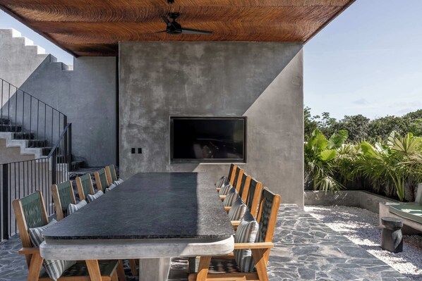 A modern outdoor dining area with a long table and chairs, a built-in TV, and a staircase leading to an upper level.