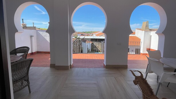 Une terrasse couverte avec vue sur les bâtiments, une chaise en osier, une table et une chèvre.