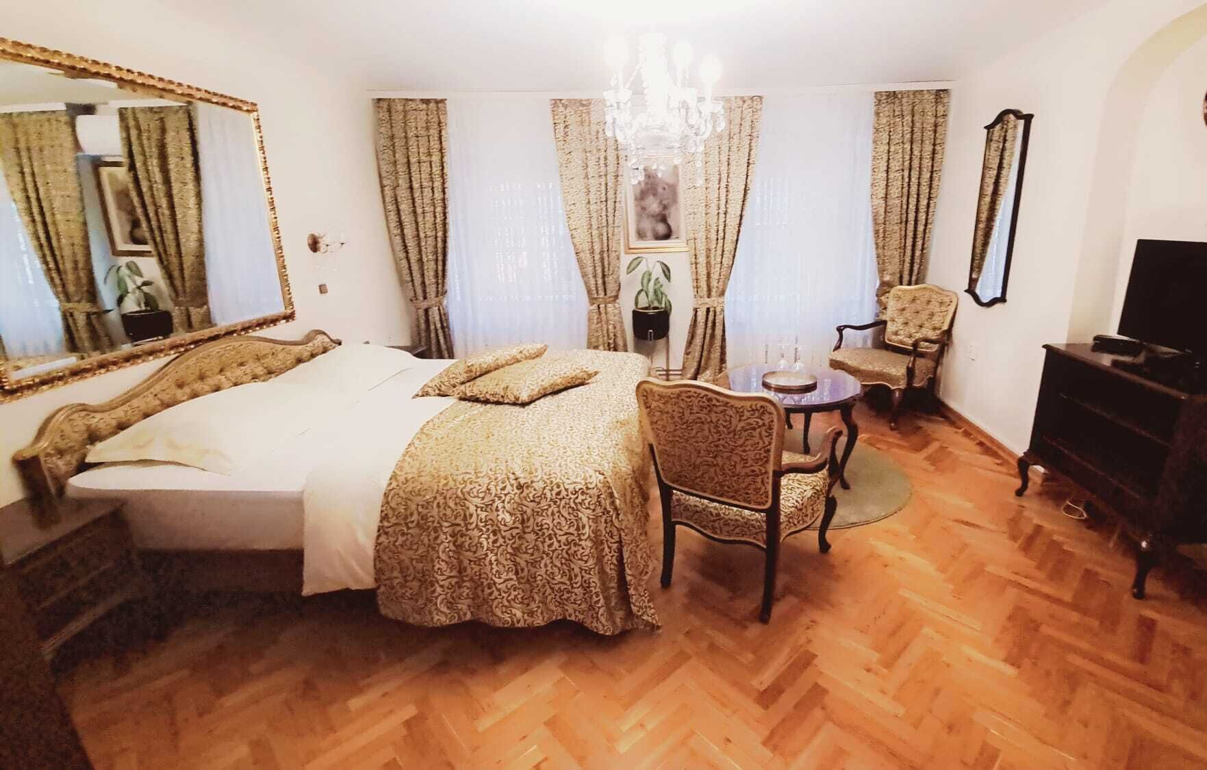 A hotel room with a large bed, a sitting area with a chair and a small table, a television, and a mirror.