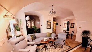 Een woonkamer met een bank, fauteuils, een salontafel en een kroonluchter. Op de achtergrond een eethoek met een tafel en stoelen.