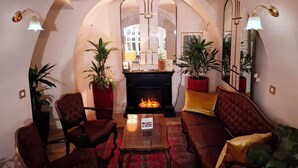 Een gezellige woonkamer met een open haard, twee fauteuils, een bank en potplanten.