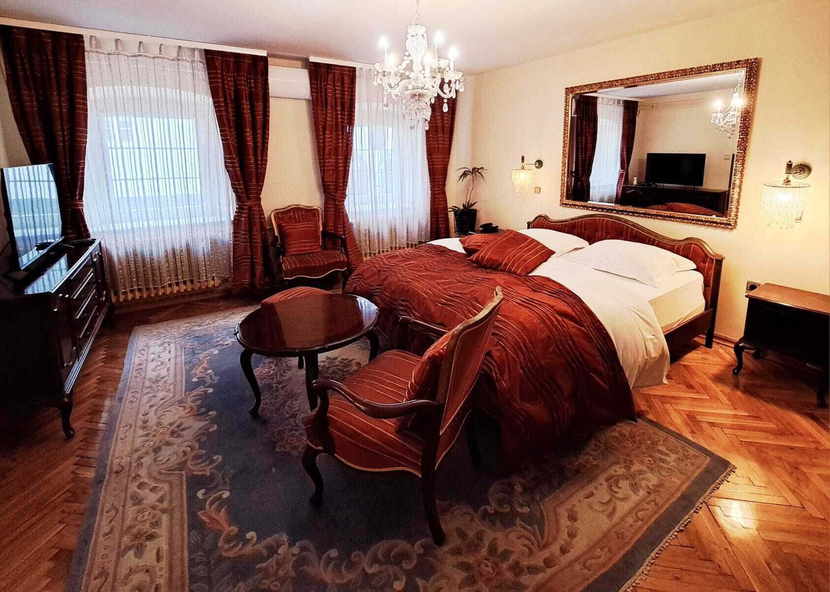 Habitación de hotel con una cama grande, una zona de estar con una mesa y sillas, un televisor, un espejo y una araña.