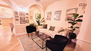 Een ruime woonkamer met een bank, twee fauteuils, een salontafel en een vloerkleed. Er hangen verschillende ingelijste foto’s aan de muren en een kroonluchter aan het plafond.