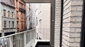 Uma varanda estreita com guarda-corpo de metal, soalho em azulejos e vista para edifícios vizinhos.
