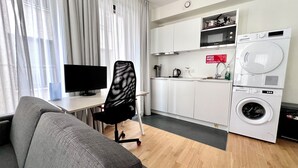 Uma sala de estar moderna com um sofá cinzento, uma cadeira de escritório preta, uma televisão de tela plana e uma área de cozinha com máquina de lavar.