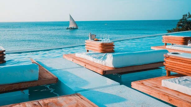 A poolside area with wooden loungers and a sailboat in the background. - Le Saint Tropez (Kidoti)