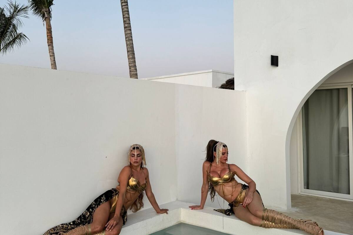 Deux femmes, vêtues de bikinis dorés, sont assises au bord d’une piscine, avec des palmiers en arrière-plan.