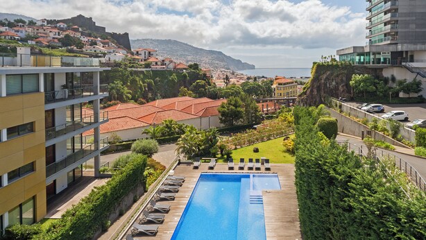 A swimming pool with lounge chairs, surrounded by greenery and overlooking a coastal town.
