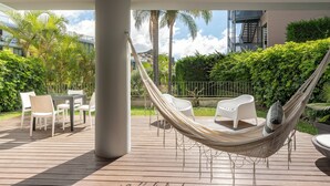 A hammock on a wooden deck with white chairs and a table, surrounded by greenery and a building in the background.