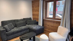 A modern living room with a sectional sofa, a white armchair, a small black table, and a window with a view of a building.