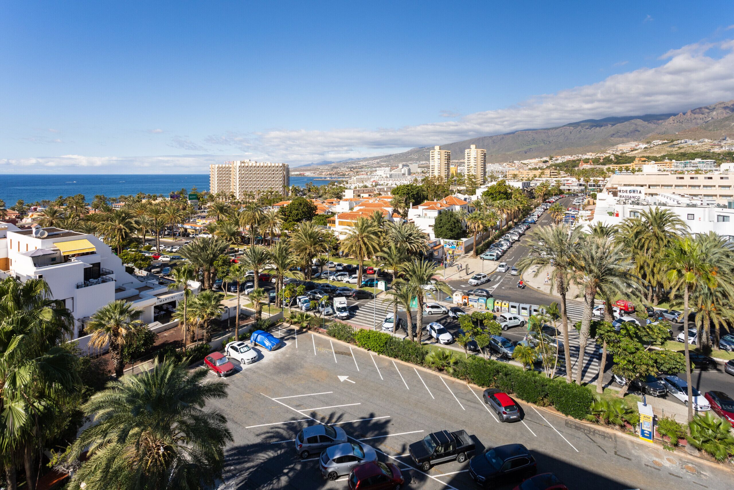 Un parking rempli de voitures, entouré de palmiers et d’immeubles.